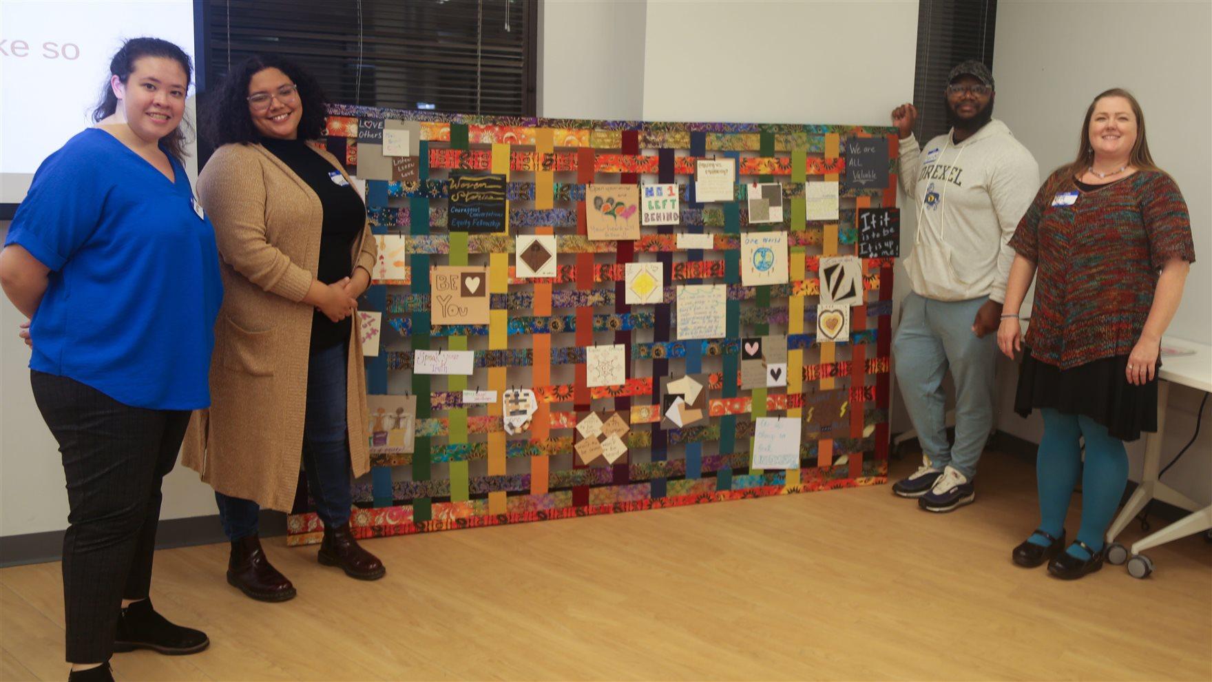 Four people stand in front of a quilt created by people who participated in the first Courageous Conversation