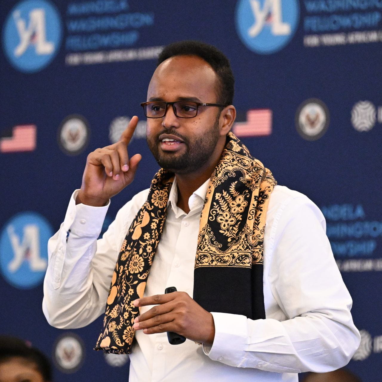 A man in a white shirt and a scarf holds a microphone in one hand while pointing up with a finger on the other hand.
