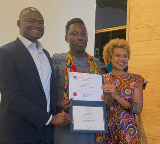 A man and a woman standing up surround a young man holding a diploma.