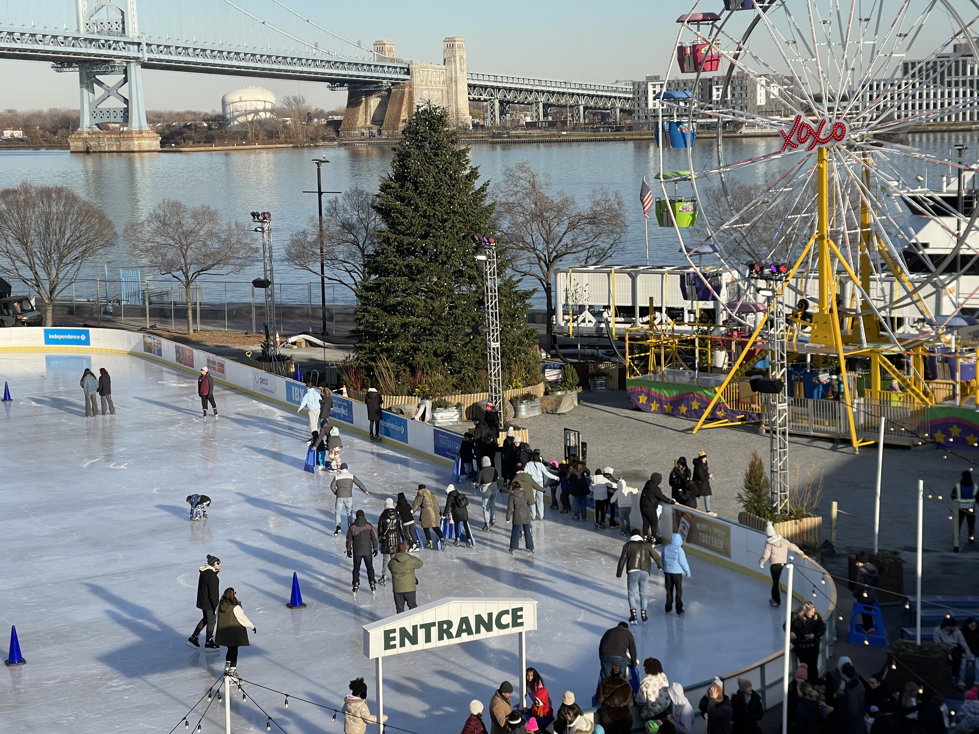 People ice skating outside on a rink next to a ferris wheel on the Delaware River.