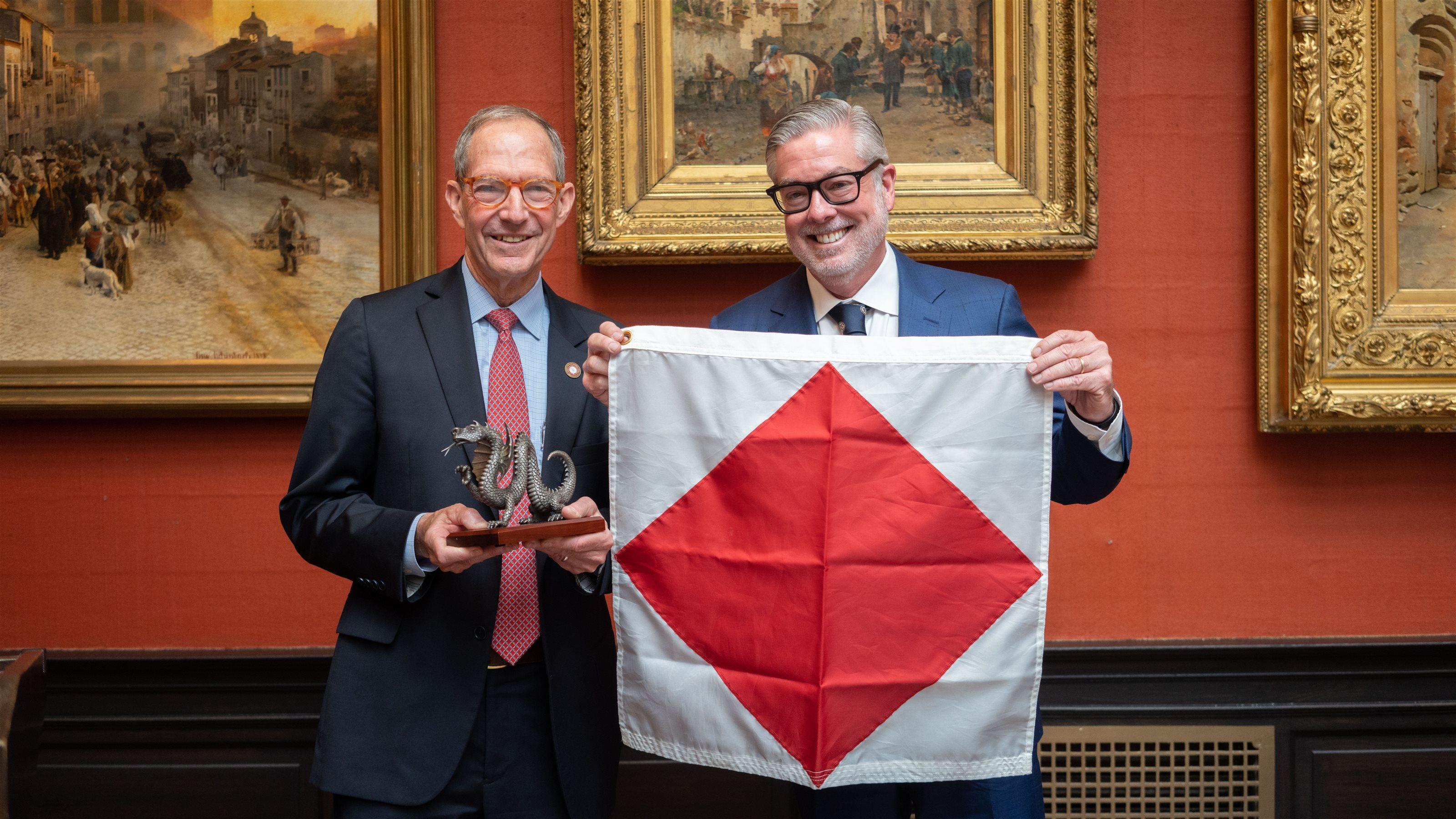 Salus University President Michael Mittelman (left) and Drexel University President John Fry standing next to each other in A.J. Drexel Picture Gallery. Michael is holding Drexel’s Mario the dragon figurine while John holds Salus’ flag (white background with red diamond in the center).